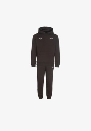 Melns kapuciņsvīteris un atbilstošas sporta bikses ar mazām baltām teksta zīmēm uz krūtīm un augšstilbiem.