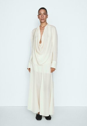 Vrouw staat tegen een witte muur, draagt een losse, crèmekleurige gedrapeerde blouse en broek met wijde pijpen met zwarte schoenen, kijkt recht vooruit.