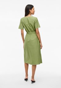 Femme de dos, portant une robe ceinturée olive à longueur genou avec des manches courtes et des mules à talons noirs sur fond blanc.