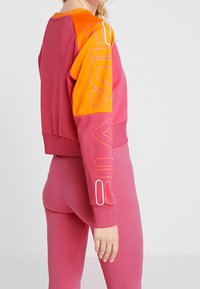 Kvinna klädd i en långärmad tröja i rosa och orange med abstrakta linjemönster på ärmarna, tillsammans med rosa leggings.