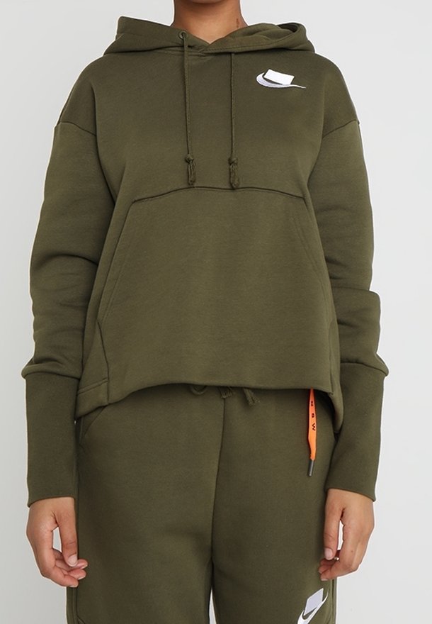 Personne portant un sweat à capuche court vert olive et un pantalon de survêtement assorti avec des logos Nike blancs et orange visibles.