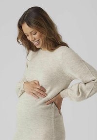 Pull en tricot beige clair avec de longues manches bouffantes et un col rond. Texture lisse, silhouette ajustée. Mains reposant sur un ventre arrondi.