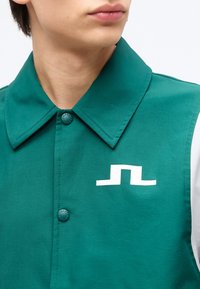 Mlada oseba, oblečena v zeleno srajco z ovratnikom, z belim abstraktnim logotipom na prsih in zelenimi gumbi z blagovno znamko.