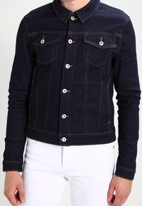 Person klädd i en mörkblå denimjacka med vita knappar och sömmar, i kombination med vita byxor, visad från halsen till höfterna.