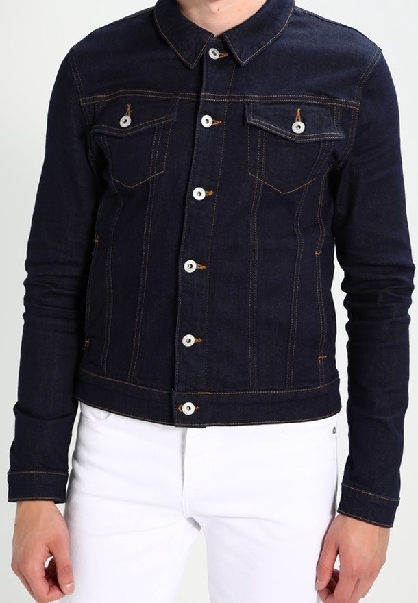 Person klädd i en mörkblå denimjacka med vita knappar och sömmar, i kombination med vita byxor, visad från halsen till höfterna.