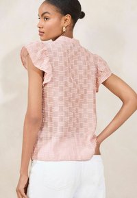 Blusa trasparente rosa con maniche corte a volant e motivo testurizzato. Il retro presenta una finitura liscia, indossata con pantaloni bianchi.