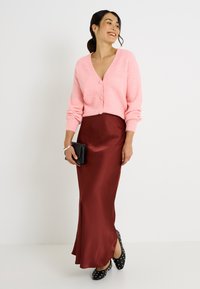 Cárdigan rosa de punto con cierre de botones, combinado con una falda ajustada de satén burdeos y zapatos planos negros con lunares blancos, sosteniendo un bolso de mano negro.