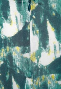 Gros plan sur un motif de tissu abstrait avec des taches vertes, jaunes et blanches, une couture verticale visible et une tirette de fermeture éclair vert foncé.
