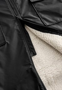 Black outerwear jacket with a zipper, featuring a soft, cream-colored shearling lining and a textured exterior.