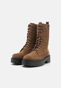 Bottines en suède marron avec un lacet sur le devant, fermeture éclair sur le côté et semelle en caoutchouc noire épaisse. Éléments décoratifs en œillets métalliques et accents cousus.
