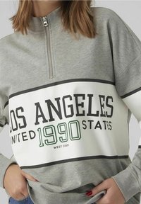 Grijze rits sweatshirt met een witte streep met "LOS ANGELES UNITED STATES 1990" in vetgedrukte zwarte en groene letters, zachte textuur.
