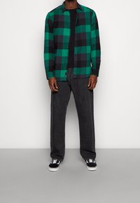 Hombre con camisa de cuadros verde y negro sobre camiseta negra, vaqueros gris oscuro y zapatillas negras y blancas de pie contra una pared blanca.
