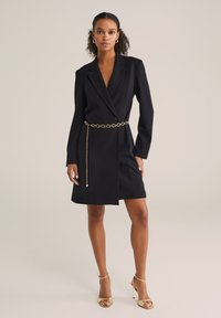 Rochie de blazer neagră cu o croială ajustată, având mâneci lungi, decolteu adânc în V și un accent cu curea din lanț auriu. Textură netedă și lungime până la genunchi.