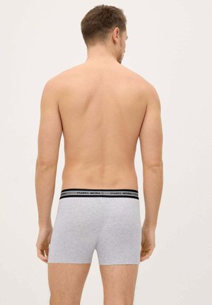 Hombre de espaldas a la cámara, vistiendo bóxers grises con cintura negra y la marca "Ysabel Mora" sobre un fondo liso.