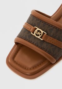 Brun slidesandal med en bred, mønstret overdel med en guldfarvet logoaccent. Blød tekstur med firkantet tådesign og syningsdetaljer.