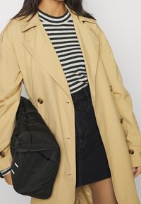 Trench-coat beige avec de gros boutons, porté sur un chemisier rayé noir et blanc et une jupe en denim noir, tenant un sac noir matelassé.