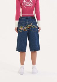 Denim shorts in dark wash, featuring colorful dragon embroidery on back pockets. Straight-leg design with cuffed hems.