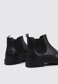 Botas Chelsea de cuero negro con paneles elásticos en los lados, tiradores y suelas de goma texturizadas. Diseño limpio sin patrones visibles.
