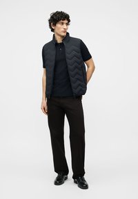 Άνδρας με σγουρά μαλλιά, φοράει μαύρο γιλέκο με ραφές πάνω από μαύρο πόλο, μαύρο παντελόνι και μαύρα δερμάτινα παπούτσια, στέκεται με τα χέρια πίσω από την πλάτη.