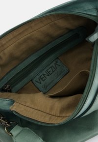 Interieur van een groene echt leren handtas, met een bruine stoffen voering, een ritsvak en een label met "VENEZIA" in reliëf.