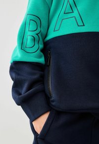 Pullover i teal og marineblå med markante sorte bogstaver "A" og "B", justerbar lynlås-lomme på siden og blød stoftekstur.