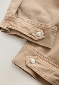 Beige denim jacket cuffs with stitched rectangular tabs secured by silver snap buttons, showing detailed fabric texture and stitching.