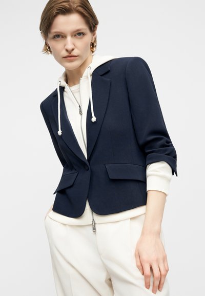 Woman wearing a navy blazer over a white zip-up hoodie and white pants, with gold hoop earrings, posing against a plain light background.