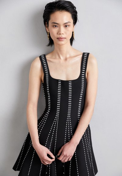 Robe noire avec un bodice ajusté ornée d'accents blancs à motifs. Encolure carrée et larges bretelles menant à une jupe évasée.