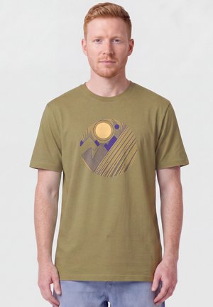 Mann mit roten Haaren, der ein olivgrünes Hemd mit einem abstrakten kreisförmigen Design mit gelben und violetten geometrischen Formen trägt.