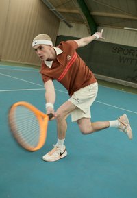 Teniso žaidėjas rudos spalvos polo marškinėliais su raudonomis juostelėmis ir balta apykakle, šviesiais šortais, mušantis teniso kamuoliuką su oranžine rakete.