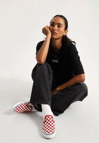 Scarpe Vans a motivi a scacchiera rosse e bianche, indossate con calze bianche, abbinate a jeans neri larghi e a una maglietta nera.