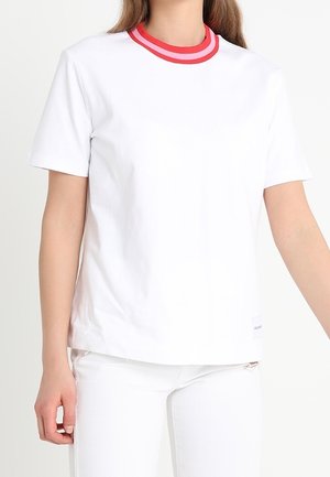 Woman wearing a white short-sleeve t-shirt with a red double-striped crew neckline and white pants against a plain background.