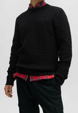 Uomo che indossa un maglione nero lavorato a maglia sopra una camicia a quadri rossa e pantaloni cargo neri, in piedi con le mani in tasca.