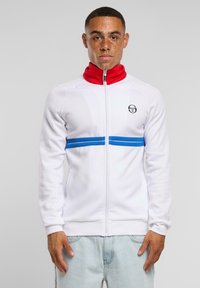 Giacca bianca con zip e colletto alto rosso, righe blu orizzontali sul petto e logo sul lato sinistro. Realizzata in tessuto liscio.
