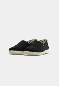 Paire de chaussures noires slip-on avec semelles blanches et doublure intérieure beige et noire à motif sur un fond uni.