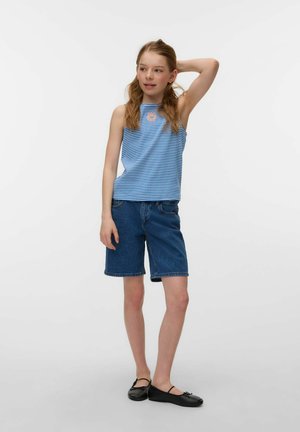 Dziewczyna ubrana w niebiesko-białą koszulkę bez rękawów w paski, krótkie dżinsowe spodenki i czarne baleriny, stoi z jedną ręką za głową.