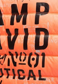 Leuchtend orangefarbene gesteppte Jacke mit auffälligen schwarzen und weißen Grafiken, einschließlich Text und nautischer Motive; glattes, glänzendes Material mit sichtbaren Nähten.