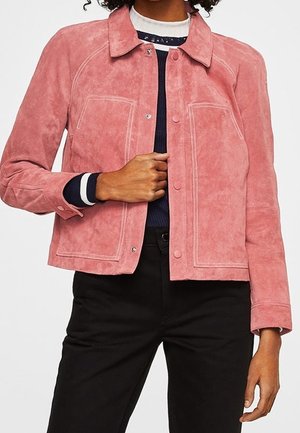 Person trägt eine pinke Wildlederjacke über einem marineblau-weiß gestreiften Pullover mit schwarzer Hose und richtet die vorderen Knöpfe der Jacke.