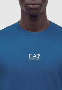 Gros plan d'une personne portant une chemise bleue avec un logo blanc EA7 Emporio Armani sur la poitrine, sur un fond uni.