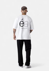 Mann mit Hals- und Arm-Tätowierungen, der schwarze Hosen, weiße Sneakers, Sonnenbrille und ein weißes T-Shirt mit schwarzem Text und Grafik auf dem Rücken trägt.