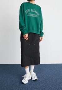 Sudadera verde con letras blancas "New Balance Athletics", combinada con una falda midi negra con una abertura frontal y detalles metálicos. Zapatillas blancas.