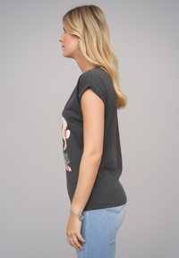 Harmaa lyhythihainen t-paita, jossa on keddareita, ja jossa on kukkakuosi. Yhdistettynä vaaleansinisisiin slim-fit-farkkuihin ja hopeiseen rannekoruun.