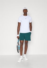 Conjunto atlético masculino com uma camisa branca de mangas curtas com um logótipo, calções verdes, ténis brancos e uma viseira, segurando uma raquete de ténis.