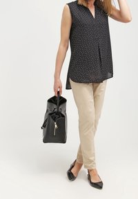 Blusa nera senza maniche con pois bianchi, pantaloni beige, borsa nera con zip, e ballerine nere a punta, in piedi su uno sfondo chiaro.