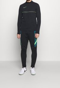 Musta urheilupaita, jossa on puolivetoketju ja vihreät korosteraidat, yhdistetty mustiin joggereihin, joissa on vihreät ja turkoosit sivupaneelit. Valkoiset lenkkitossut.