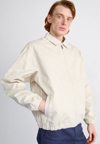 Lys beige jakke med krave, elastiske ærmer og en afslappet pasform. Har sidelommer og en diskret logo på brystet.