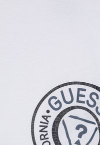 Camiseta de algodón blanca con un gran gráfico circular que presenta un texto quebrado en negro y gris que dice "GUESS" y un diseño de signo de interrogación.
