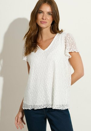 Frau trägt eine weiße Spitzenbluse mit kurzen Ärmeln und V-Ausschnitt und dunkelblaue Jeans, steht vor einem schlichten hellen Hintergrund.