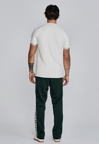 Camiseta blanca con mangas acentuadas en verde, combinada con pantalones jogger verde oscuro que presentan un patrón floral vertical en el costado. Ajuste casual.
