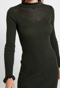 Ribbat, långärmad klänning i mörkgrön med en hög, fläktad halsringning och en meshpaneldetalj över bröstet.